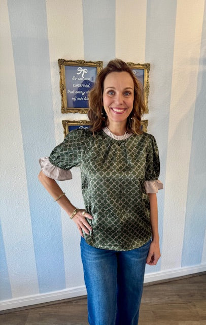 Woman wearing a patterned blouse with ruffled sleeves in front of a wall with striped wallpaper.