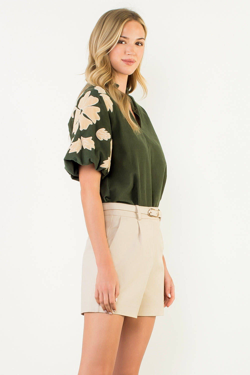 Woman wearing a green blouse with floral sleeves and beige shorts on a white background