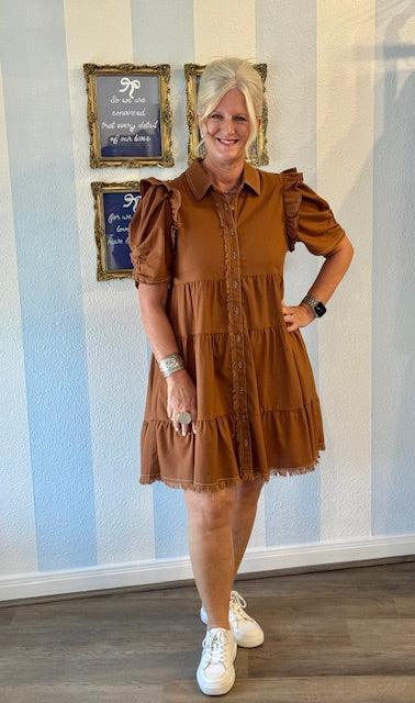 Woman wearing caramel brown tiered button-front dress with puff sleeves, ruffle details, and frayed hem, styled with sneakers and silver jewelry.