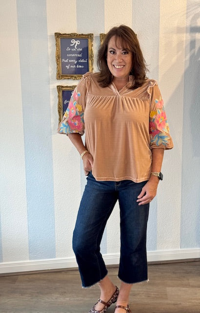 Woman wearing champagne velvet top with colorful embroidered 3/4 sleeves, ruffled neckline, and relaxed fit, styled with dark jeans.