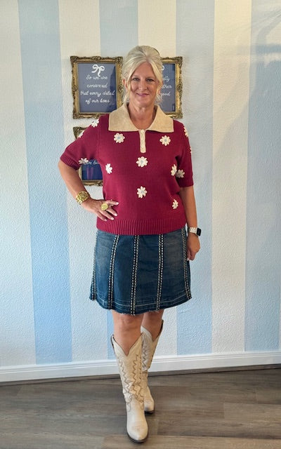 Woman wearing a red floral sweater, blue skirt, and white boots in a room with striped wallpaper.