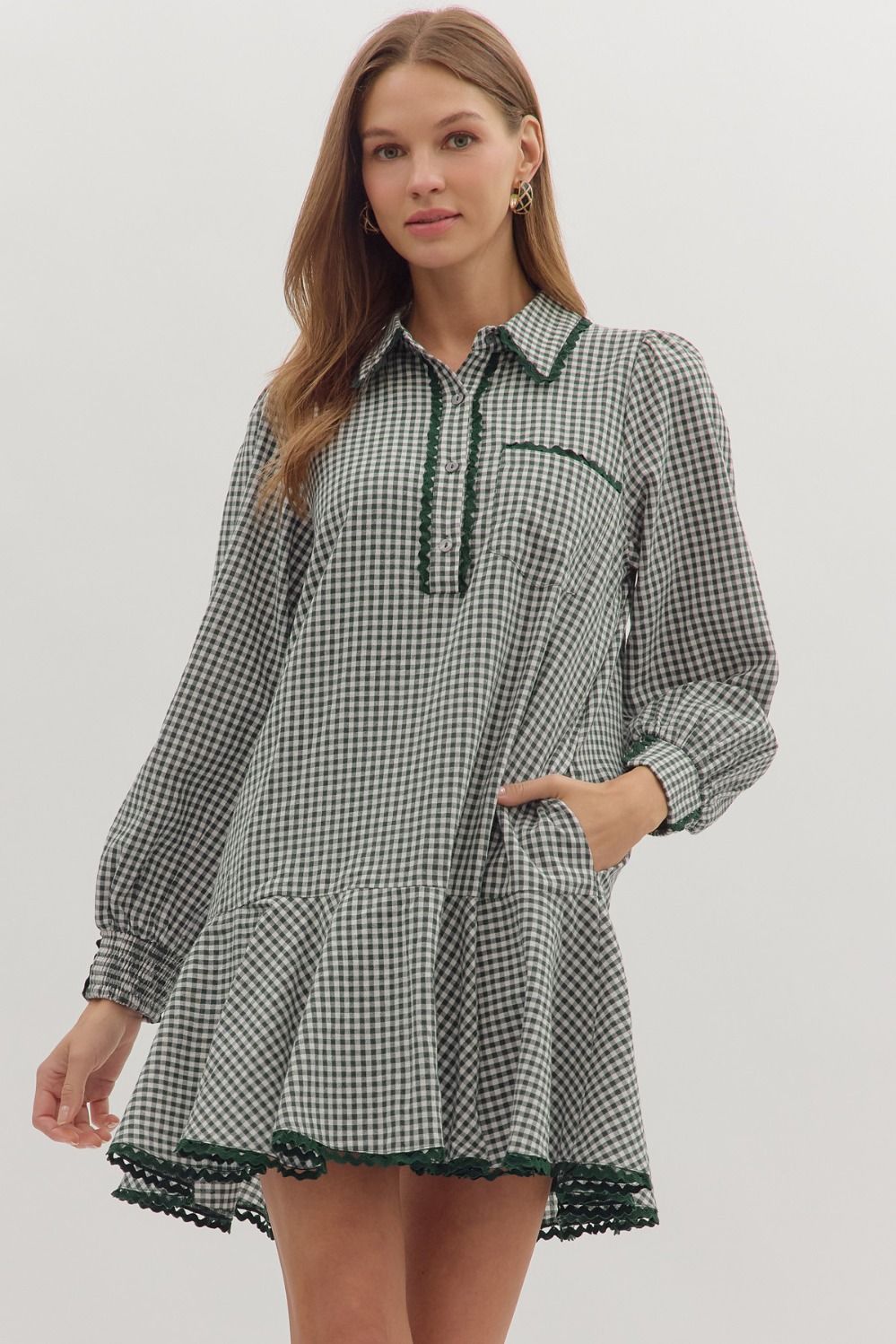 Woman wearing a green and white gingham long-sleeve mini dress with a collared neckline, ric-rac trim details, tiered hem, and side pockets, standing against a light background