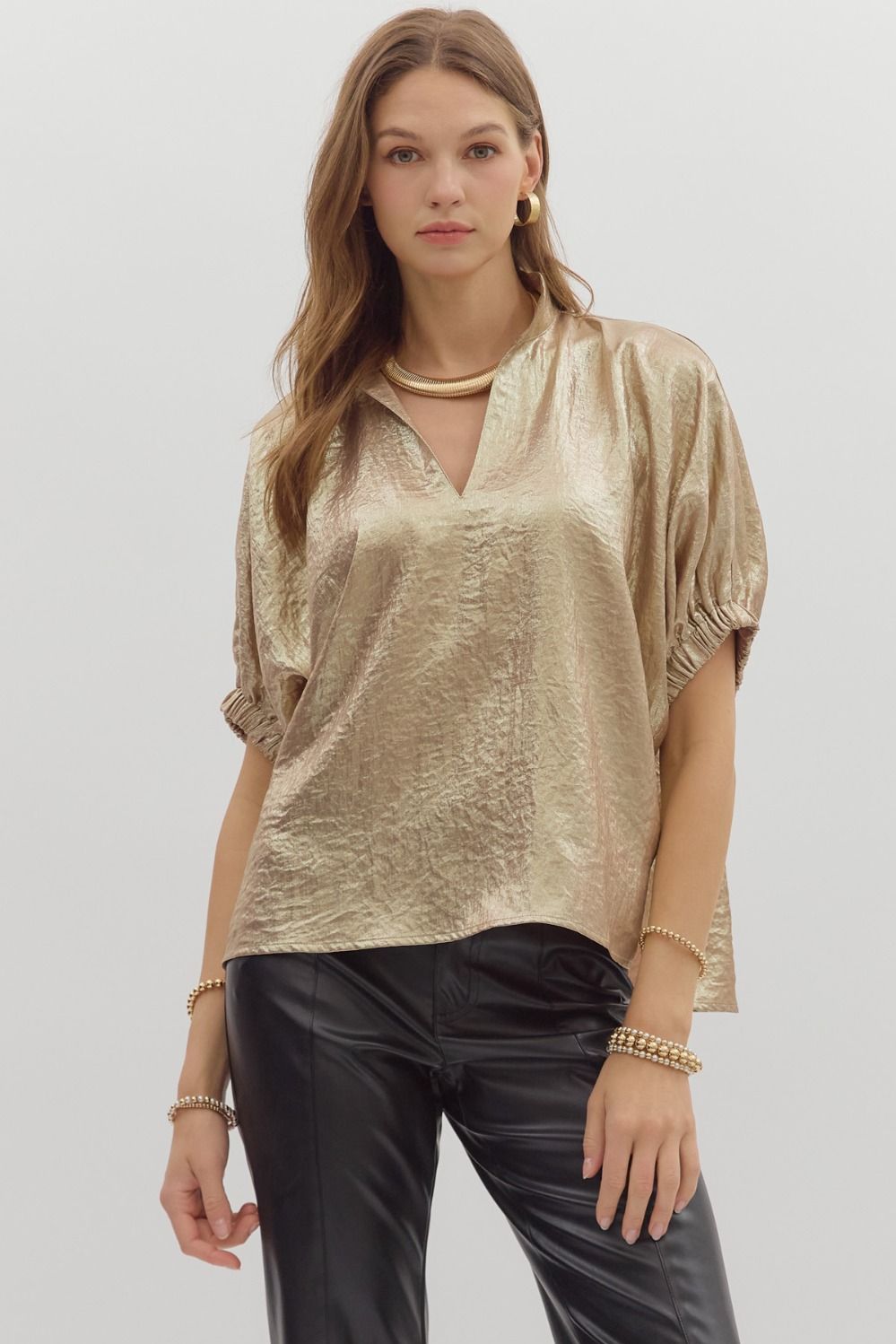 Woman wearing a metallic gold crinkle-textured blouse with a V-neckline and gathered short sleeves, paired with black leather pants against a light background