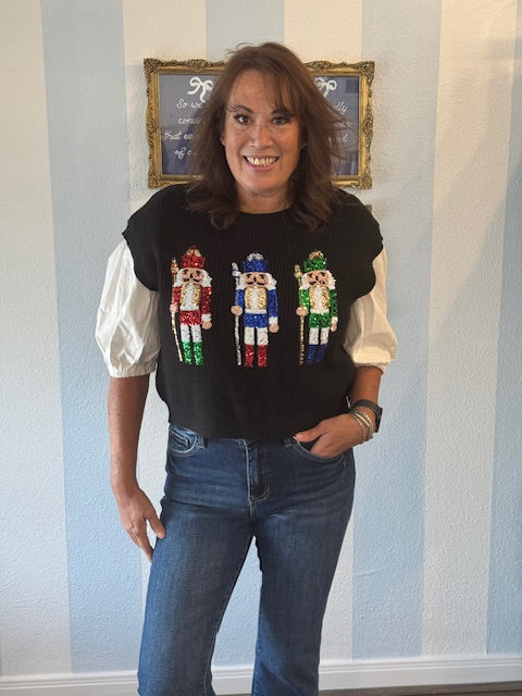 Black sweater vest featuring colorful sequin nutcracker designs across the front, styled over a puff-sleeve blouse with denim jeans for a festive holiday look.
