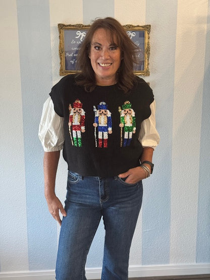 Black sweater vest featuring colorful sequin nutcracker designs across the front, styled over a puff-sleeve blouse with denim jeans for a festive holiday look.