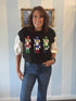 Black sweater vest featuring colorful sequin nutcracker designs across the front, styled over a puff-sleeve blouse with denim jeans for a festive holiday look.