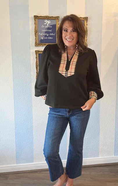 Woman wearing a black blouse with a plaid pattern and blue jeans standing in front of a striped wall.