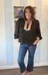 Woman wearing a black blouse with a plaid pattern and blue jeans standing in front of a striped wall.