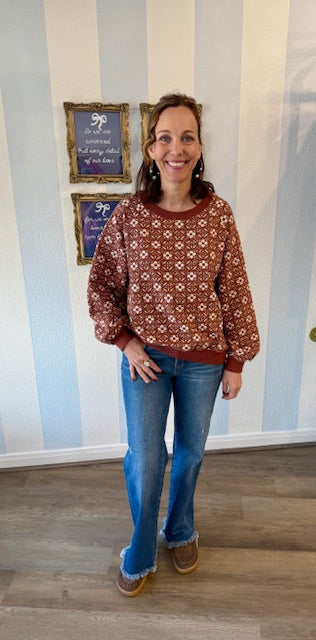 Woman wearing rust pullover with cream geometric print, long sleeves, and banded cuffs, styled with jeans and tan mules.