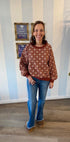 Woman wearing rust pullover with cream geometric print, long sleeves, and banded cuffs, styled with jeans and tan mules.
