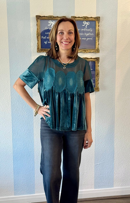 Woman wearing a teal blouse with a patterned design standing in front of a wall with framed pictures.