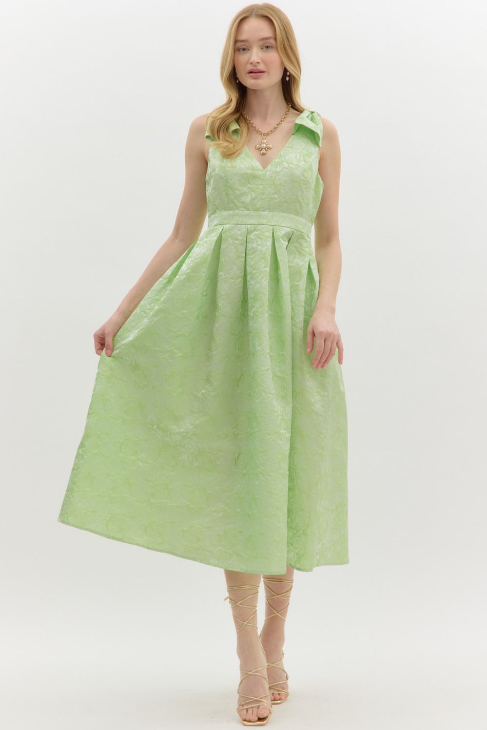 A dress featuring a delicate floral textured fabric in a soft lime green hue. Designed with a flattering V-neckline, pleated skirt, and an oversized bow accent at the shoulder. Also features side pockets and a back zipper.