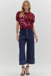 Woman wearing a maroon sweater with pink floral design and blue pants on a white background